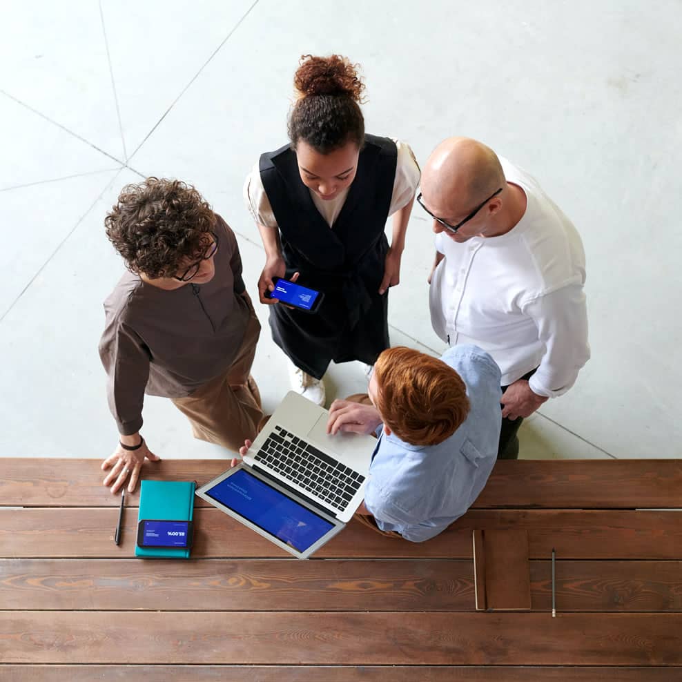 Four colleagues collaborating using a laptop and smartphone around a wooden table. Four colleagues collaborating using a laptop and smartphone around a wooden table.